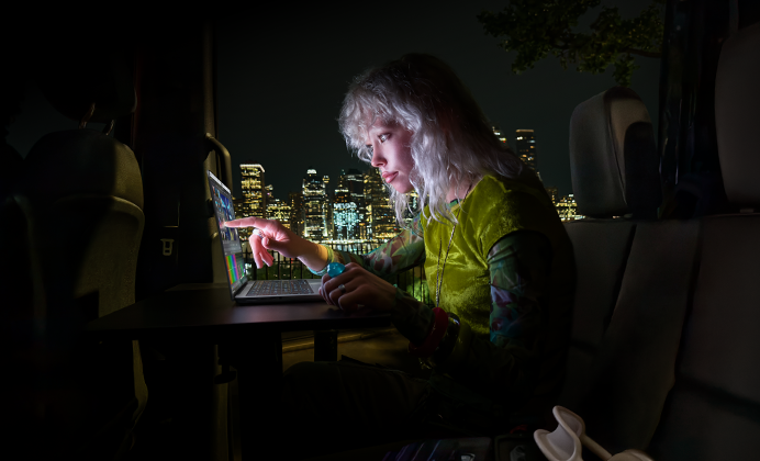 Eine Person sitzt auf dem Rücksitz eines Vans, durch die geöffnete Beifahrertür ist die Skyline einer Stadt bei Nacht zu sehen, ein MacBook Pro mit PROLED XDR Display auf einem Schreibtisch, die Person wird vom Bildschirm beleuchtet, während sie das Touch Feature nutzt, um ein Video in DaVinci Resolve Studio zu bearbeiten